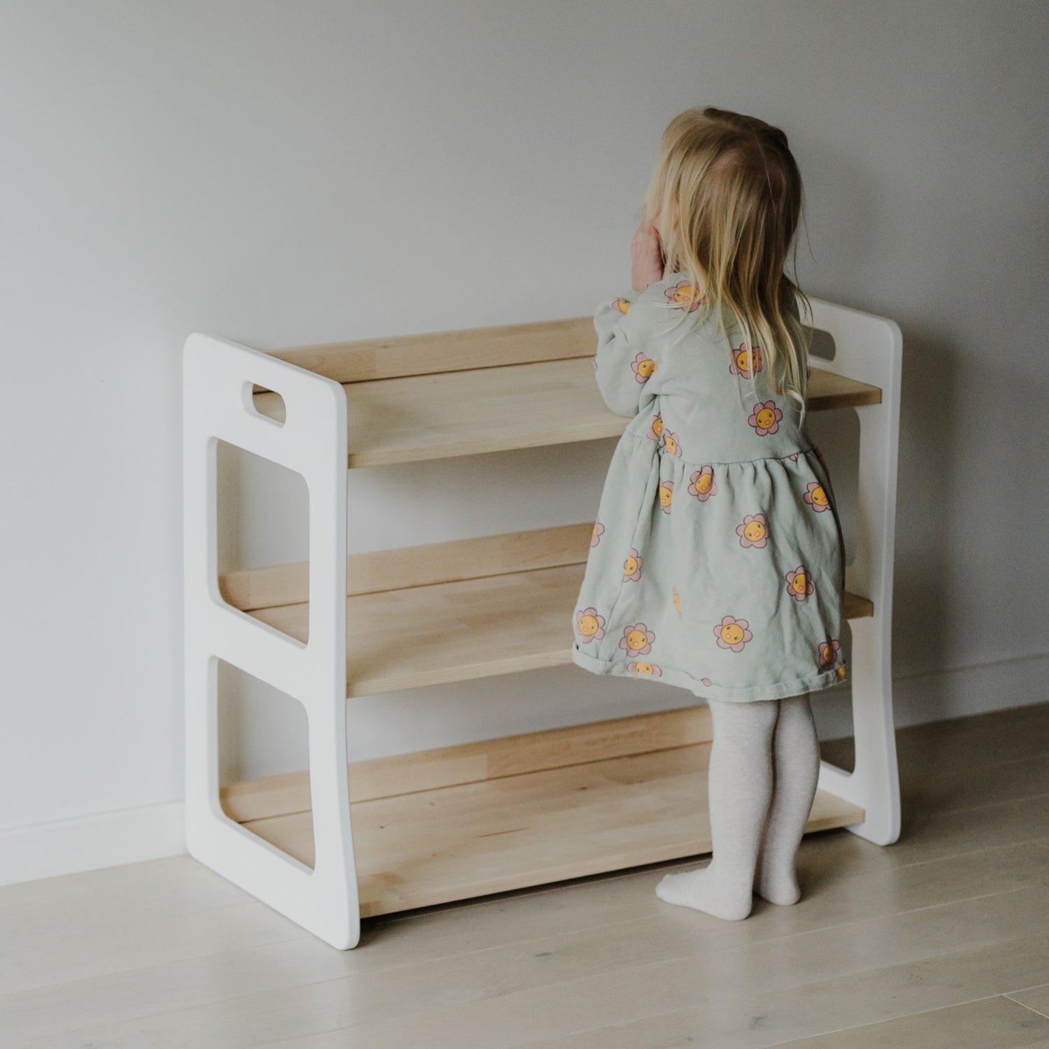 Ein Kleinkind steht vor einem leeren Montessori Spielzeugregal der HARMONIA Kollektion, welches vor einer weißen Wand auf Laminatboden steht. Das Regal ist offen gestaltet und auf perfekter Greifhöhe des Kindes gebaut.