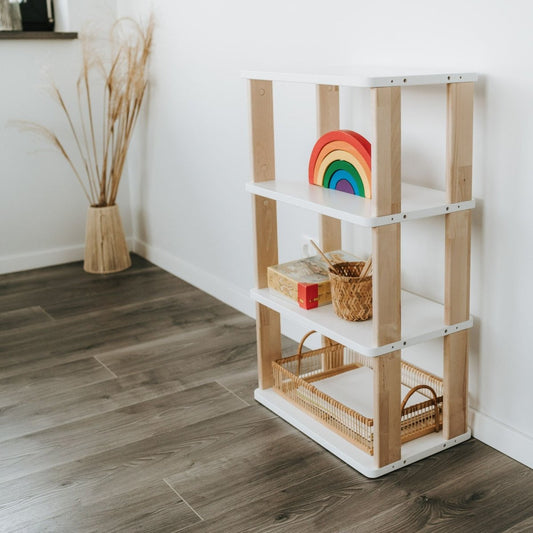 Das kleine Montessori Regal der MODURA Kollektion mit vier weiß lackierten Ablagen & mit Klaröl veredelten Seitenteilen steht vor einer Wand. Ein Spielbogen, ein Korb & weitere Produkte sind in den Regalfächern harmonisch angeordnet.