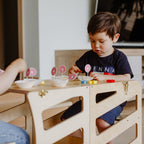 var:klarlack-lernturm / var:klarlack-lernturm-rutsche / var:klarlack-lernturm-hocker / var:klarlack-lernturm-rutsche-hocker / Detailaufnahme des zum Kindertisc umgebauten Lernturmes im Streifen-Design in Klarlack und eines Kindes, welches auf der zugehörigen Sitzbank sitzt und mit Knete spielt.