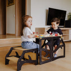 var:schwarz-lernturm-hocker / var:schwarz-lernturm-rutsche-hocker / Zwei Geschwisterkinder sitzen gemeinsam am zum Kindertisch umgebauten schwarzen Lernturm im Baum-Design. Sie scheinen zu spielen und dabei Muffins zu essen.