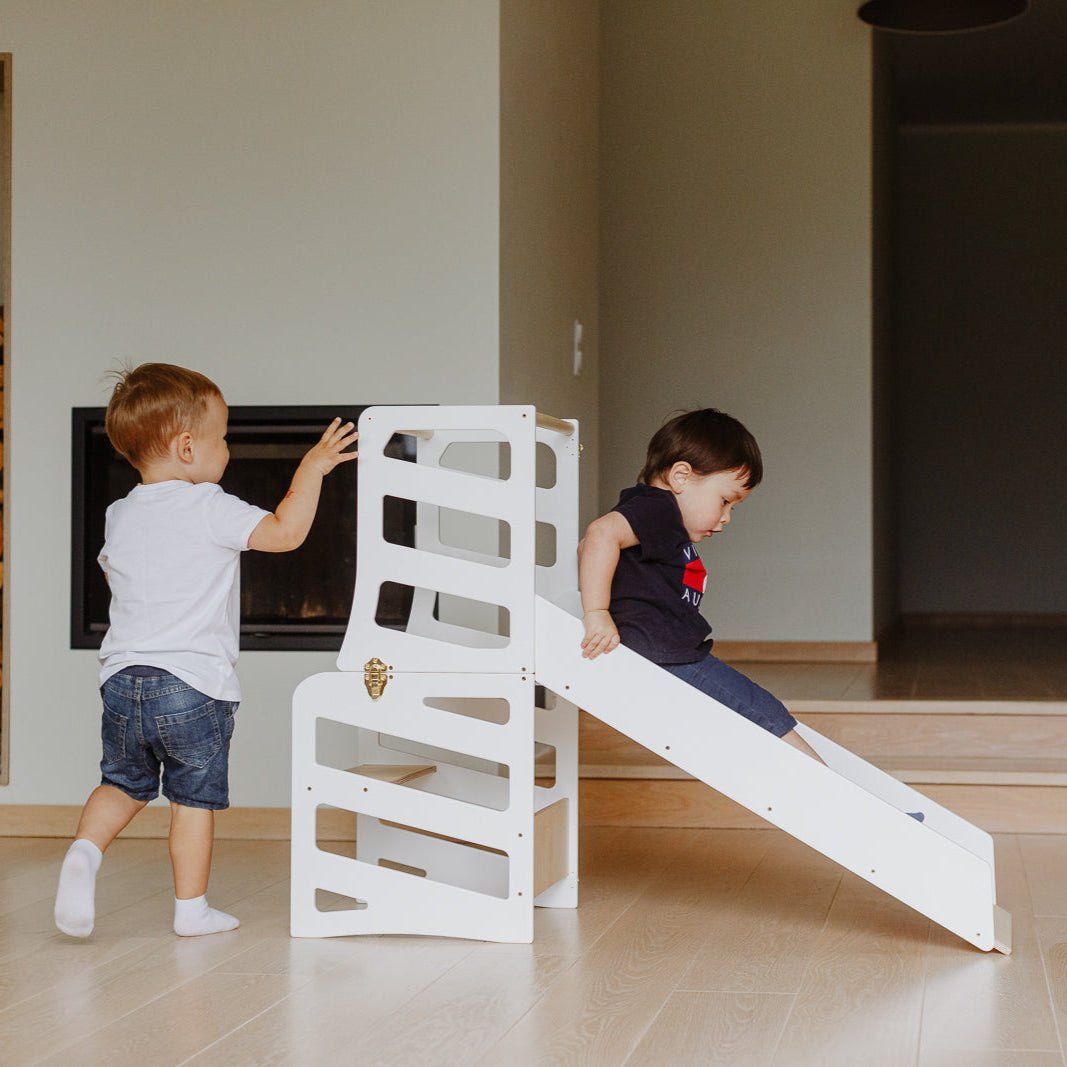 var:weiß-lernturm-rutsche / var:weiß-lernturm-rutsche-hocker / Ein Kind nutzt die Rutsche des weißen Lernturms im Streifen-Design. Ein anderes Kind möchte danach ebenfalls rutschen.