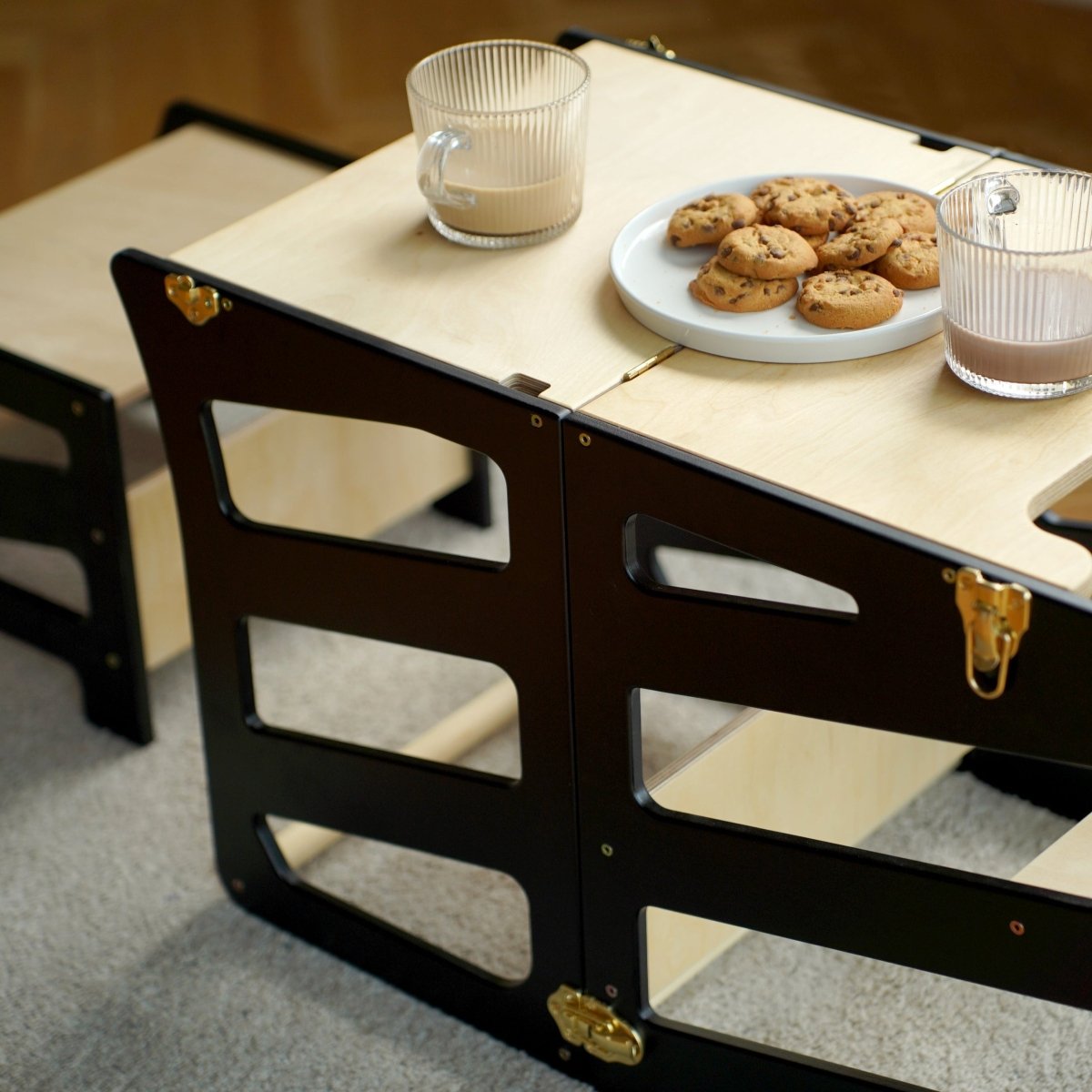 var:schwarz-lernturm-hocker / var:schwarz-lernturm-rutsche-hocker / Seitenansicht der Tischfunktion des schwarzen Lernturmes mit Extra-Hocker im Streifen-Design. Auf der Tischplatte stehen zwei Gläser mit Milch sowie ein Teller mit Keksen.