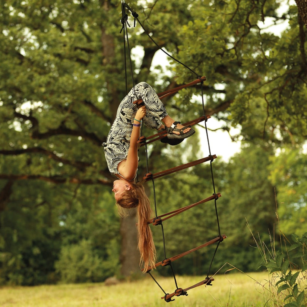 Ein Kind hängt mit den Beinen an den obersten Holzsprossen der Strickleiter Größe L in braun kopfüber herunter und hält sich an den robusten Seilen fest.
