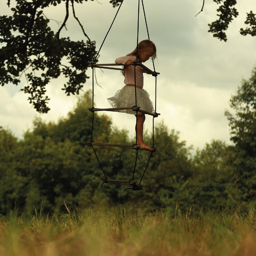 Ein Kleinkind steht fokussiert mit den Füßen auf unteren Holzsprossen der Strickleiter in grau  und hält sich mit den Händen an den oberen Sprossen fest. 