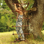 Ein Mädchen hält sich an den oberen Klettersprossen der braunen Strickleiter in der Größe L fest, welche an einem Baum befestigt ist. Das Kind steht innerhalb der Dreieck-Strickleiter aus Holz auf den untersten Sprossen.