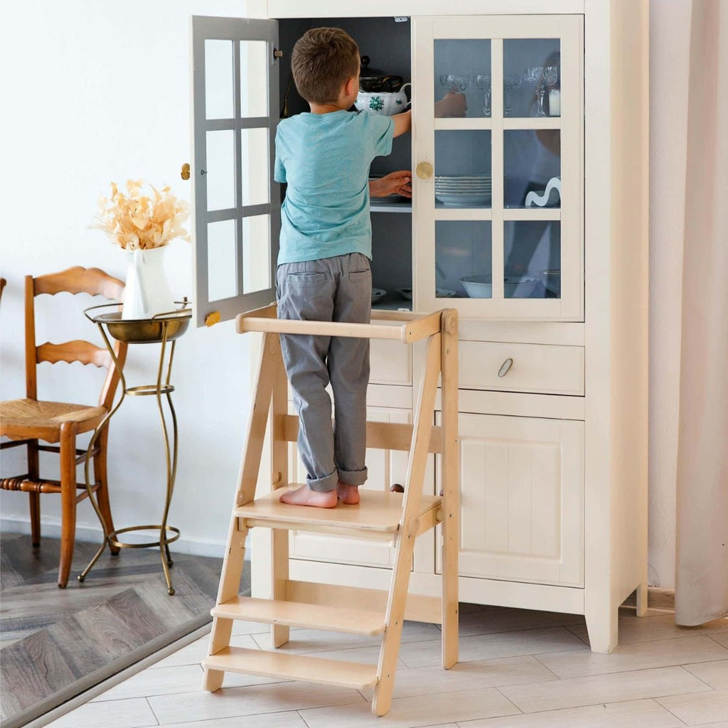 Ein Kind steht auf der Plattform des klappbaren Lernturmes und holt Geschirr aus einem Schrank.