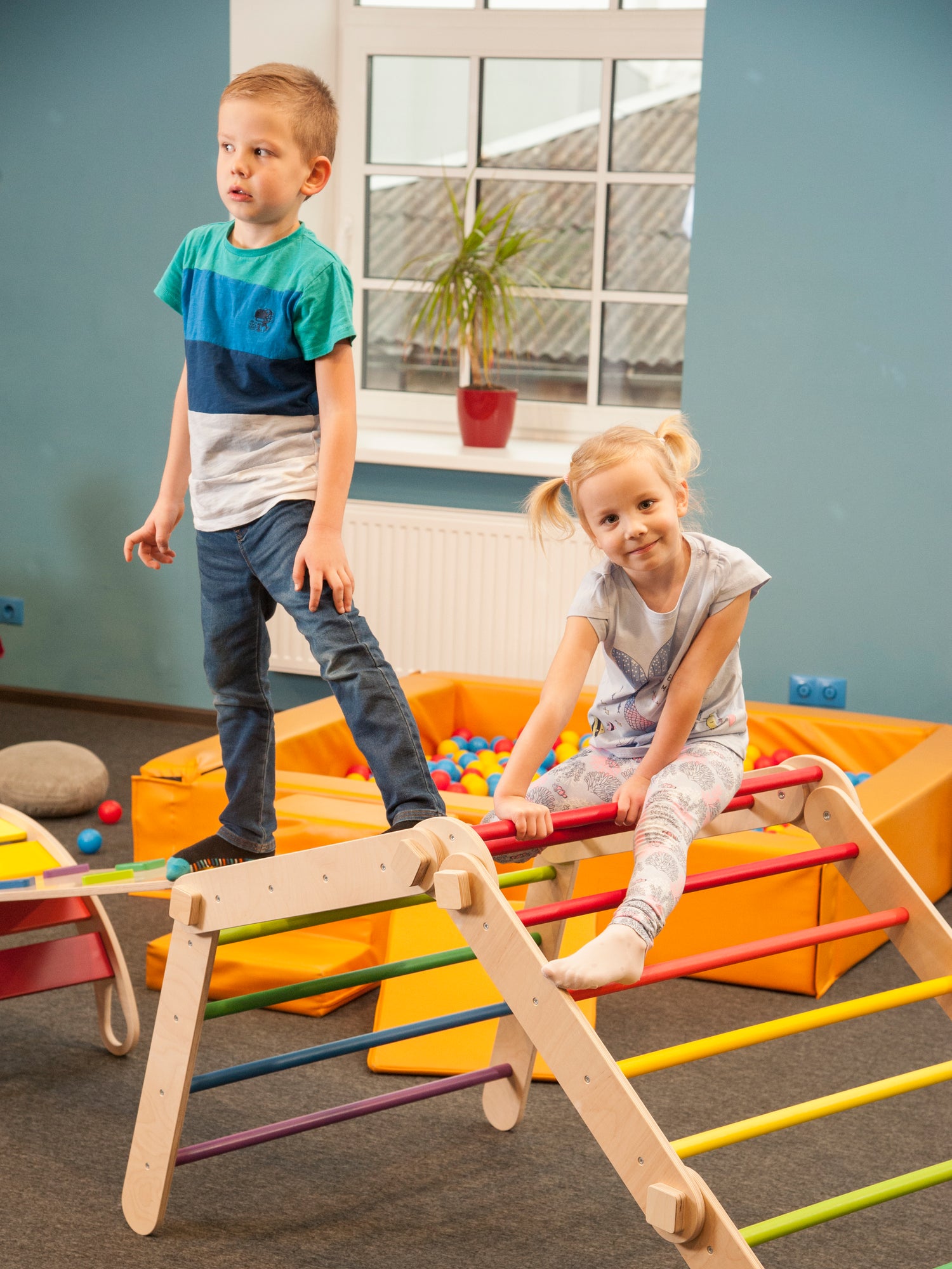 Hier sind ein Junge und ein Mädchen zu sehen, wie sie auf einem Indoor-Klettergerüst, bestehend aus einem verstellbaren Kletterdreieck mit Rutsche sowie einem Kletterbogen spielen.