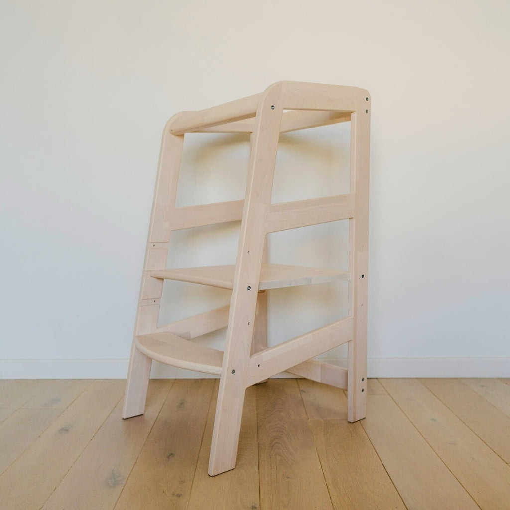 Der Lernturm aus Massivholz ohne montiertem Rausfallschutz steht vor einer weißen Wand auf einem Holzboden mit Dielen.