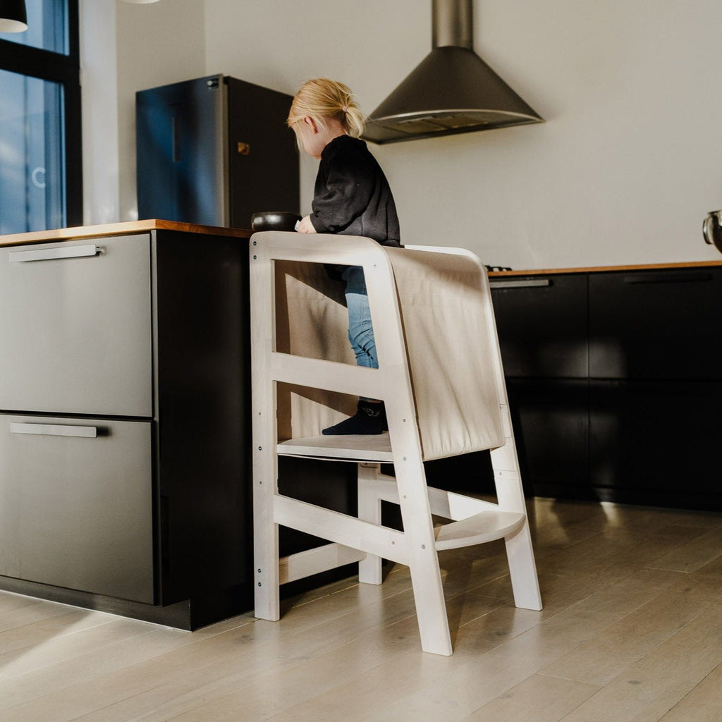 Ein Kleinkind steht auf der Plattform des Massivholz-Lernturmes mit Rausfallschutz vor einer schwarzen Küchentheke.
