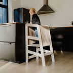 Ein Kleinkind steht auf der Plattform des Massivholz-Lernturmes mit Rausfallschutz vor einer schwarzen Küchentheke.