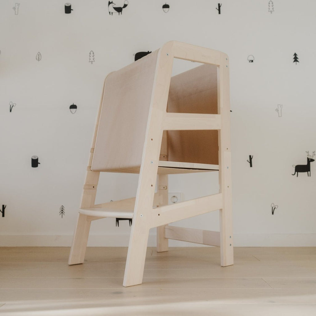 Der Lernturm mit Rausfallschutz steht vor einer Wand in einem Kinderzimmer mittig im Bild.