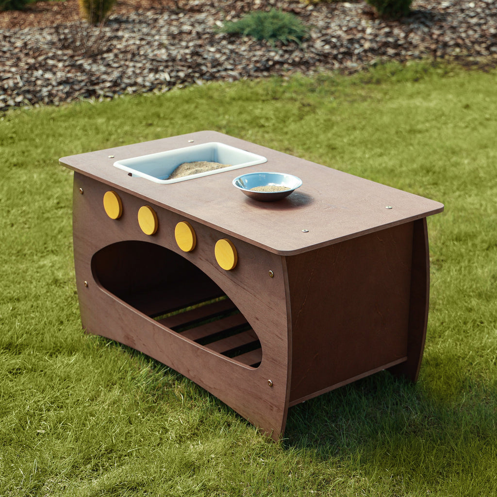 Die Outdoor Matschküche aus der seitlichen Ansicht auf einer grünen Wiese. Der inkludierte Behälter ist mit Sand befüllt und eine Metallschüssel steht auf der Ablage.