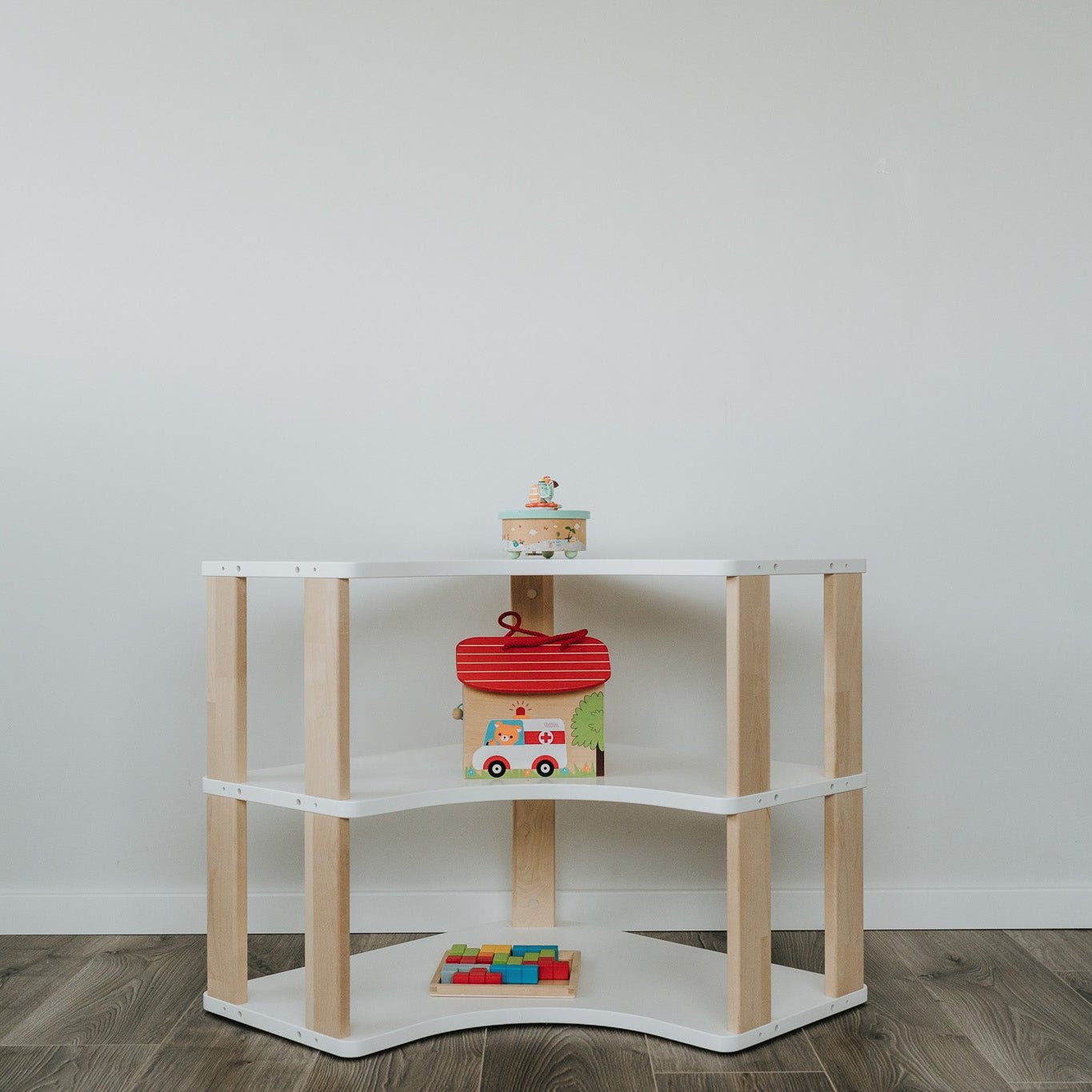 das Eckregal steht mittig im Kinderzimmer vor einer weißen Wand. Ausführung mit 3 Ablagen in weiß + klares Öl.