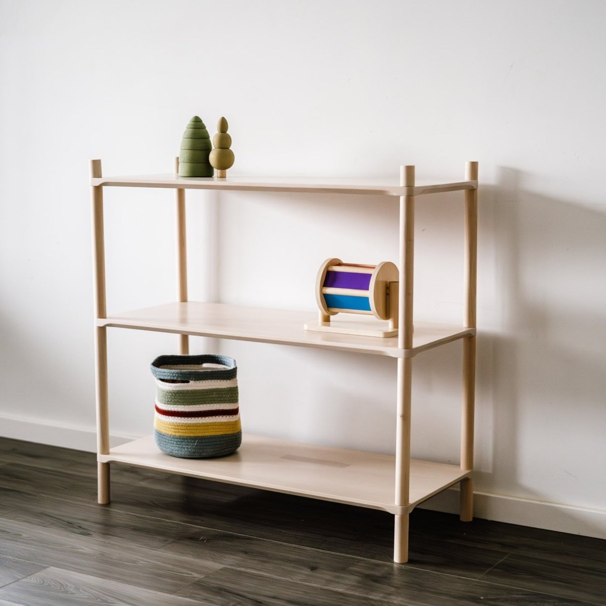 Die Midi-Variante des ISKORA Holzregales für Kinderzimmer & Wohnräume mit drei Regalböden angelehnt an einer weißen Wand. Spielzeug und ein Korb aus Stoff sind darin angeordnet.