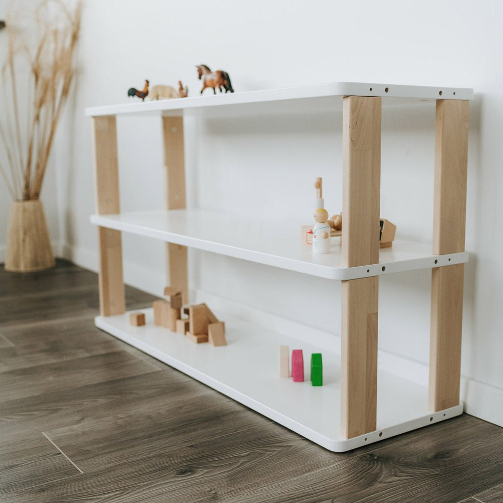 Das modulare Kinderregal nach Montessori der MODURA Kollektion mit drei weiß lackierten Ablagen und geölten Seitenplanken steht vor einer weißen Wand in einem Kinderzimmer. Auf den Ablagen sind Spielzeuge & Dekoartikel angeordnet.