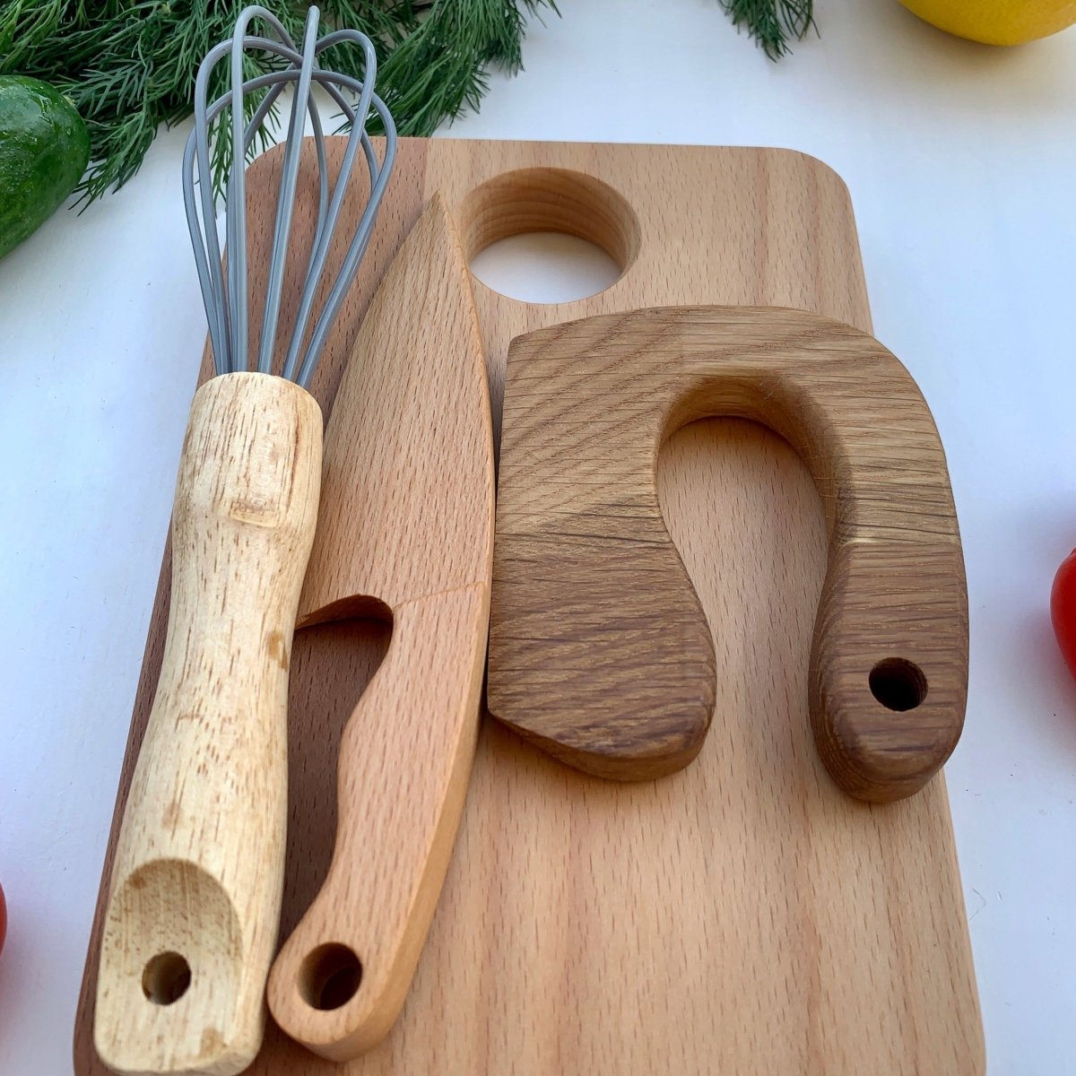 Nahaufnahme eines Montessori Küchensets. Man sieht ein Frühstücksbrett auf dem ein Schneidemesser, ein Kindermesser und ein Schneebesen nach Montessori liegen.