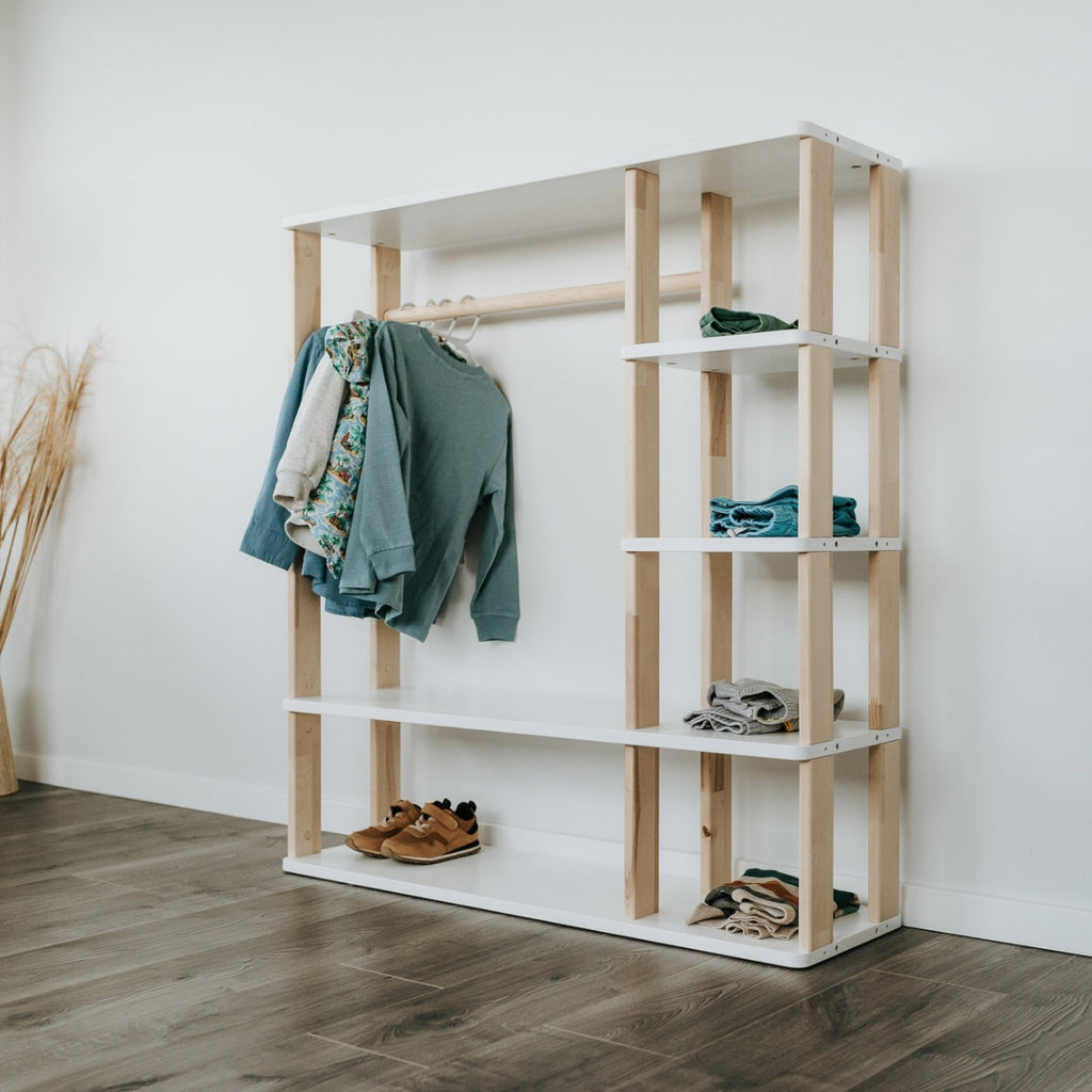 Unser Montessori Kleiderschrank mit weißen Oberflächen steht vor einer weißen Wand. In den offenen Fächern sind Klamotten und Schuhe verstaut und an der Kleiderstange hängen Pullover und Hemden an Kleiderbügeln.