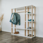 Unser Montessori Kleiderschrank mit weißen Oberflächen steht vor einer weißen Wand. In den offenen Fächern sind Klamotten und Schuhe verstaut und an der Kleiderstange hängen Pullover und Hemden an Kleiderbügeln.