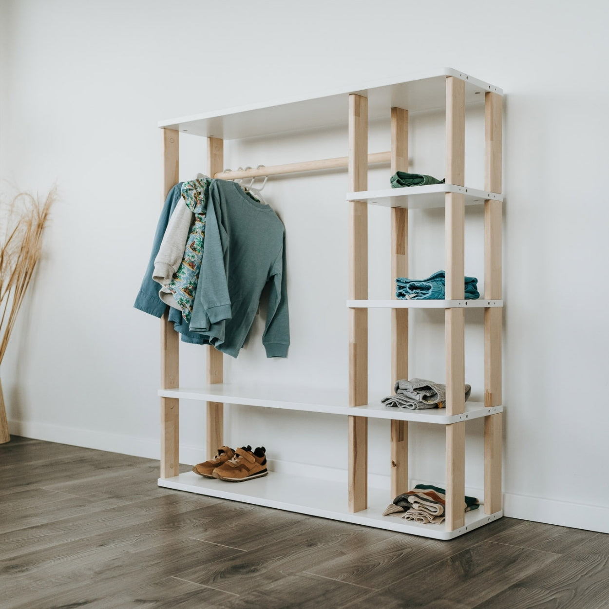Unser Montessori Kleiderschrank mit weißen Oberflächen steht vor einer weißen Wand. In den offenen Fächern sind Klamotten und Schuhe verstaut und an der Kleiderstange hängen Pullover und Hemden an Kleiderbügeln.