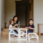 var:weiß-lernturm-hocker / var:weiß-lernturm-rutsche-hocker / Zwei geschwisterkinder sitzen gemeinsam am zum Kindertisch umgebauten weißen Lernturm im Baum-Design mit Extra-Hocker. Sie spielen und die Mutter hilft dem einen Kind dabei ein Problem zu lösen. 