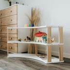 Das Montessori-inspirierte Bogenregal für Kinder der MODURA Kollektion mit drei weißen Ablagen & geölten Seitenplanken steht neben einer Kommode vor einer Wand im Kinderzimmer. In den Regalfächern sind verschiedene Figuren aus Holz angeordnet.